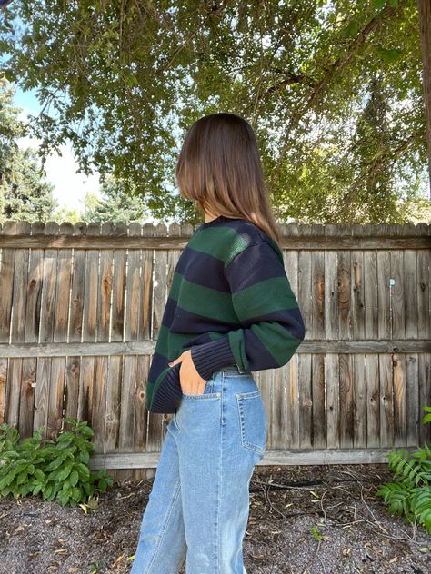 Woman standing in front of a weathered brown fence, wearing light wash baggy jeans and a sweater with large green and navy blue stripes. She is not looking at the camera and is facing sideways (left). There are green trees and plants in the background and the sun is peeking through the tree leaves in the top right corner Fall Sweater And Jeans Outfit, Jean Sweater Outfit, Baggy Jumper Outfit, Jumper And Jeans Outfit, Sweater Jeans Outfit, Jeans And Sweater Outfit, Baggy Sweater Outfits, Levi Jeans Outfit, Jeans Fall Outfit