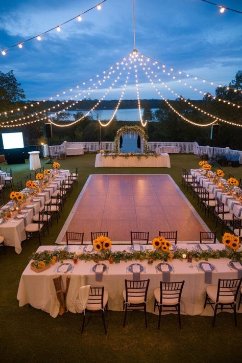 An outdoor wedding reception setup with a dance floor in the center, surrounded by tables decorated with sunflowers and greenery. String lights hang above, creating a romantic atmosphere. A scenic view of water and trees is in the background. Small Modern Wedding Ideas, Outdoor Diy Wedding Reception, Wedding Inspo Romantic Small, Wedding Ideas Minimal, Evening Outside Wedding, Cute Intimate Wedding Ideas, Classy Outdoor Wedding Reception, Outdoor Fall Wedding Ideas September, Medium Sized Wedding Ideas