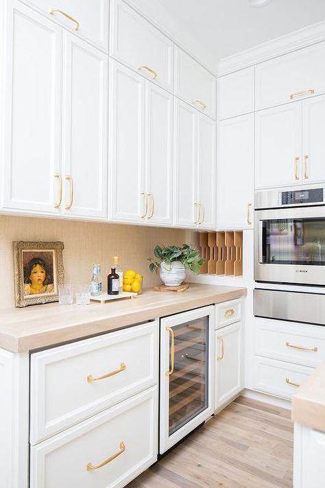 Elegant white and gold kitchen pantry boasts white shaker cabinets accented with brass pulls and holding a glass front wine fridge over a rustic wood floor and beneath a beige countertop matching a gold raffia backsplash. White And Gold Kitchen, White Kitchen Pantry, Small Kitchen Pantry, White Pantry, Rustic Wood Floors, Pantry Cabinets, White Shaker Cabinets, Wood Floors Wide Plank, Beige Kitchen