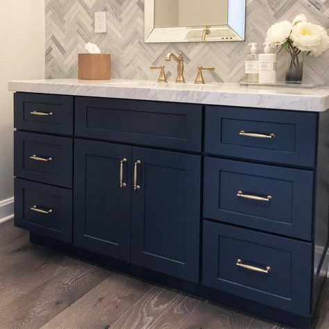 What’s not to love about this stunning powder room!? The blue vanity cabinet pairs so perfectly with the satin brass hardware. The tile wall in the chevron pattern is trendy but the neutral grey palette is very versatile. #bathroom #bathroomdesign #bathroomremodel #powderroom #interiordesign #remodel #interior #designer #interiordesigner #blue #bluecabinets #bluebathroom #homedesign Bathroom Tile Ideas With Blue Cabinets, Navy And Grey Bathroom Decor Ideas, Dark Blue Wall Bathroom, Blue Bathrooms, Navy Blue Bathrooms, Satin Brass Hardware, Wainscoting Bathroom, Gray Counter, Modern Coastal Home