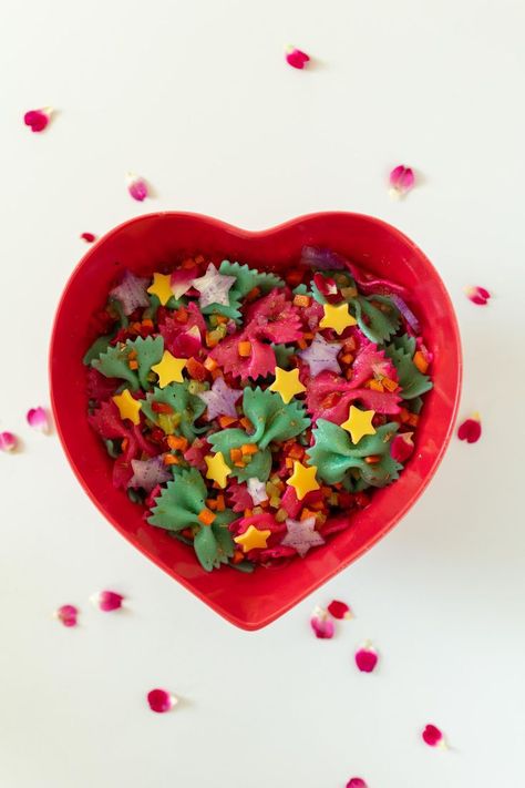 A heart shaped bowl with rainbow pasta salad inside on a table sprinkled with rose petals. The pasta shapes are bowties, and the cheese and veggie shapes are stars. Italian Bowtie Pasta, Pasta Salad With Cheese, Sailor Moon Birthday Party, Dinner Pasta Salad, Rainbow Pasta Salad, Moon Birthday Party, Salad With Cheese, Sailor Moon Birthday, Rainbow Pasta