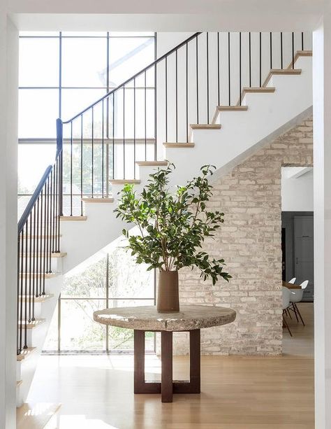 Minimalist foyer boasts a round concrete table topped with a potted plant and positioned in front of white staircase finished with an iron handrail and spindles and wood treads illuminated by natural light from a large black factory window accented with wall half covered in brick. Boho Floral Nursery, Foyer Staircase, Floral Nursery Decor, Mediterranean Style Home, Interior Finishes, Open Staircase, Mediterranean Home Decor, Take The Stairs, Mediterranean Decor