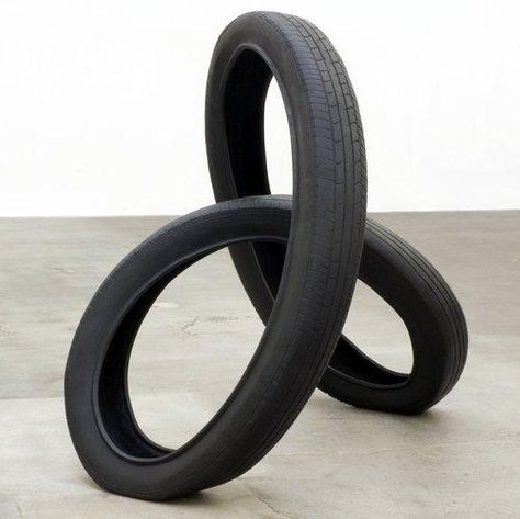 A tire, looped and twisted in a shape resembling a three-dimensional figure-eight, sits on a concrete surface. Each loop intersects with the other, forming a three-pointed structure. Sculpture Abstract, Artistic Space, Contemporary Sculpture, Sculpture Installation, Stone Rocks, The Shape, Car Art, Art Sculpture, Medium Art