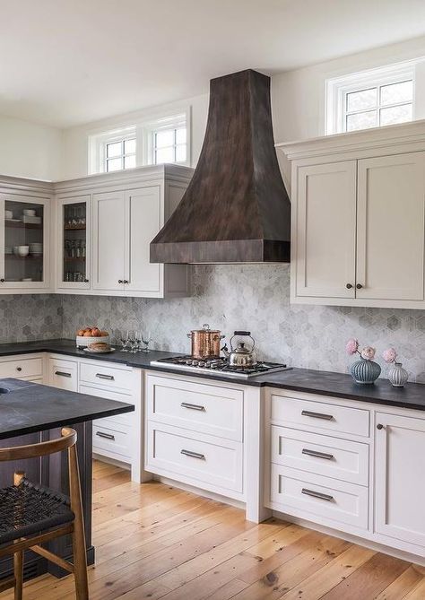 Windows are located above white shaker cabinets lined with crown molding and positioned beneath windows located on either side of a distressed black metal French range hood. Cabinet Crown Molding, French Range, Wood Floor Design, Recessed Panel Cabinets, Wood Floor Kitchen, Kitchen Windows, White Shaker Cabinets, Hood Design, Cottage Kitchens