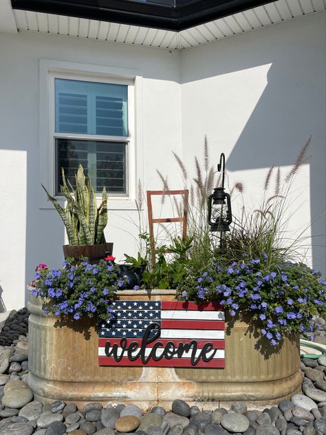 A variety of zone, sunlight, and soil plants inside a livestock tank with added visual interest such as a rustic bucket, a ceramic hen nestled in the plants, a small shepherd’s hook with an antique lantern, and a patriotic sign hanging on the front. At night, the display, which sits between the driveway and front door, is lit up by a solar spotlight. Repurposed Water Trough, Stock Tank Repurposed, Water Trough Ideas Backyards, Cow Trough Garden, Trough Flower Planters, Trough Flower Garden, Horse Trough Planter Ideas, Metal Horse Trough Planters, Old Water Trough Ideas