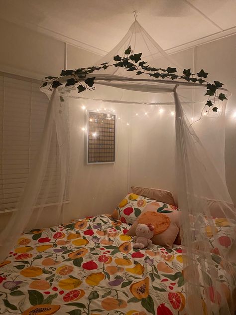 White room with queen bed. Peach themed quilt cover with a white canopy on top with vines and soft yellow lights. Pink Bed Canopy Aesthetic, Cottage Core Canopy Bed, Above Bed Canopy, Corner Bed Canopy Ideas, Bed With Canopy Aesthetic, Corner Canopy Bed, Bed Canopy Aesthetic, Corner Bed Canopy, Diy Bedroom Canopy