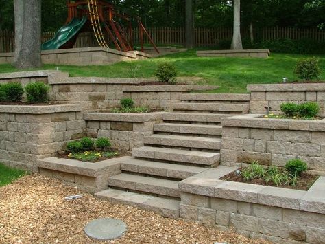 This stone walkway leads up to a stairway that matches the scene perfectly. The neutral gray stones in this setup makes the colors around it seem much more vivid and bright. Using the retainer walls on either side as planter’s boxes makes the scene uniformed without taking too much away from the natural ambience. Plants with vivid flowers would look great filling these stone garden boxes. Cinder Block Garden Wall, Terraced Patio Ideas, Retaining Wall Steps, Backyard Retaining Walls, Sloped Backyard Landscaping, Cinder Block Garden, Sloped Backyard, Landscaping Retaining Walls, Garden Stairs