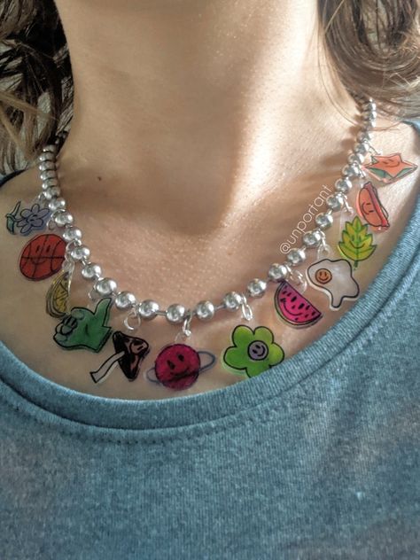 mirror selfie of a model's neck wearing a stainless steel ball chain necklace with colorful handmade small plastic charms. all charms have smiley faces and include a shooting star, hotdog, leaf, fried egg, watermelon slice, green flower, ringed striped planet, spotted mushroom, green thumbs up, lemon slice, basketball, and a blue flower with a leafy stem. source: @unportant Shrinky Dink Necklace, Diy Shrinky Dink, Shrinky Dink Art, Shrink Paper, Shrinky Dink, Fun Jewelry, Shrink Plastic, Paper Clay, Clay Ideas