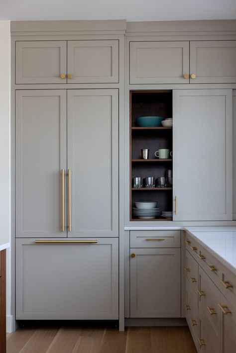 A streamlined kitchen corner boasts taupe Shaker style cabinetry with gold hardware, and chic white marble countertops. Cabinet doors slide to expose custom built-in shelves for easy organization, and the hidden refrigerator adds a sophisticated touch. Hidden Appliances, Shaker Cabinets Kitchen, Coastal Color Scheme, Yarmouth Maine, Open Shelving Kitchen, Gray Doors, Taupe Kitchen, Shaker Style Kitchen, Shelving Kitchen