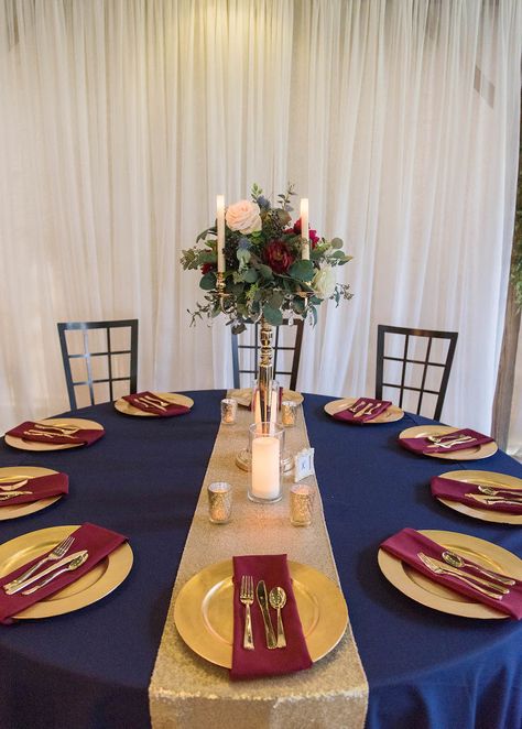 Rustic Wedding Reception Decor, Round Tables with Navy Blue Tablecloths, Gold Chargers and Burgundy Linens, Tall Silver Candlestick with Greenery, Ivory and Red Floral Bouquet Centerpiece, Gold Table Runner, Bistro Hanging Lights | Rustic Tampa Wedding Venue Kathleen's Garden Navy Blue And Burgundy Wedding Table Decor, Navy Blue And Burgundy Wedding Reception Tables, Wedding Reception Table Linens, Navy Wedding Decorations, Navy And Burgundy Wedding, Blue Wedding Receptions, Bouquet Centerpiece, Gold Chargers, Rustic Wedding Reception