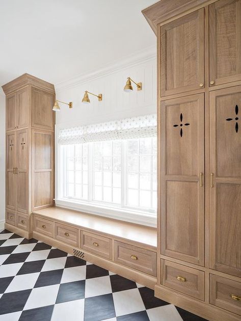 Chic black and white harlequin floor tiles end at a light brown oak mudroom bench boasting drawers accented with brass cup pulls and fitted beneath a window dressed in a white roman shade lit by brass sconces. Mudroom Lockers With Window, Master Closet Entry From Bathroom, Mud Room Windows, Blue Gray Mudroom Cabinets, Wainscoting In Entryway, Sunroom Floor, Harlequin Floors, Mudroom Cabinets, Mudroom Lockers