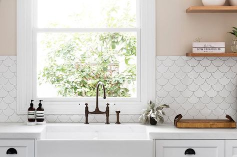 Cream kitchen walls accented with Tabarka Studio Artisan Terracotta Fan Tiles frame a window positioned above a farmhouse sink finished with a bronze faucet fixed to a honed white marble countertop located under stacked floating wooden shelves and complementing white shaker cabinets donning oil rubbed bronze cup pulls. Cream Kitchen Walls, Fish Scale Tile Backsplash, White Fish Scale Tile, Kitchen Wall Tiles Backsplash, Kitchen Wall Design, Scale Tile, Redo Kitchen Cabinets, Trendy Farmhouse Kitchen, White Farmhouse Sink