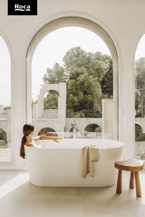 In front of an arched window is a long, oval bathtub. Sat in it is a woman of fair skintone with a brown ponytail - the ponytail falls on the outside of the bath tub. In the centre of the tub is a silver faucet. A beige towel is hanging from the side of the bathtub. To the right is a small, oddly shaped wooden stool. Roca Bathroom, Oval Bath, Neutral Bathroom, Bathtub Drain, Bathroom Inspo, Bathtubs, Bathroom Inspiration, Chrome Finish, Free Standing