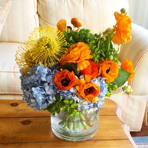 Springtime floral arrangement with blue hydrangea, orange ranunculus, yellow pincushion protea and euphorbia Blue And Orange Flower Arrangements, Blue White Orange Flower Arrangements, Orange And Blue Flower Arrangements, Wedding Colora, Blue Orange Yellow Centerpiece, Blue Orange Floral Arrangement, Blue Yellow Orange Flower Arrangements, Green Event Decor, Blue Orange Yellow Floral Arrangement