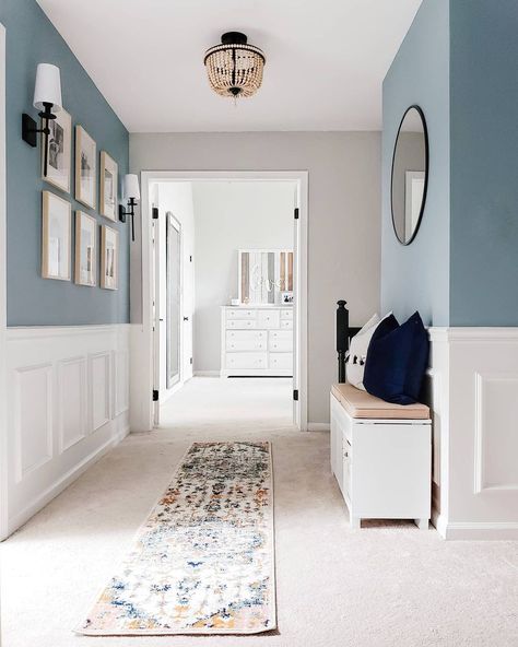 This hallway boasts beautiful color contrasts with white wainscoting fitted along a muted blue wall. A white bench seated under a black circle mirror creates a place to sit and admire a light wood frame gallery wall on the other side. Decorate A Narrow Hallway, Decorating A Narrow Hallway, Modern Wainscoting Ideas, Hallway Mudroom, Blue Hallway, White Hallway, Hallway Paint, Window Mudroom, Hallway Colours