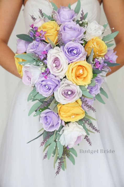 This lovely assortment of lavender, wisteria and yellow roses is bound to turn heads on your wedding day. This lovely cascading brides’ bouquet from the is an absolutely breathtaking arrangement for any bride to be, in any season. Bright greenery and lavender and lilac accents with pearls complete the look and add a delicate realism to this beautiful bouquet. This bouquet is 10” wide, 18” long, and is crafted by our in-house florists without using a foam base to anchor the flowers to the bouquet Yellow Roses And Lavender Bouquet, Garden Theme Bouquet, Purple And Yellow Bridal Bouquet, Purple And Yellow Wedding Ideas, Wisteria Flower Bouquet, Lavender And Yellow Wedding Theme, Rapunzel Flower Bouquet, Purple And Yellow Wedding Flowers, Yellow And Purple Wedding Theme