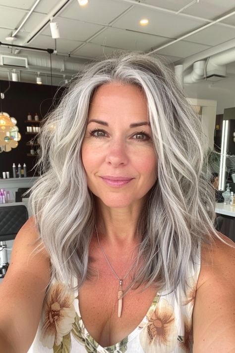 Smiling woman with long wavy gray hair, wearing a floral top, in a modern salon setting. Platinum Grey Blending, Gradual Grey Hair Aging Gracefully, Two Tone Gray Hair, Womens Salt And Pepper Hair, Grey Blonde Curly Hair, Grey Blended Highlights, Grey Hair With Shadow Root, Blended White Hair, Gray Grow Out With Highlights