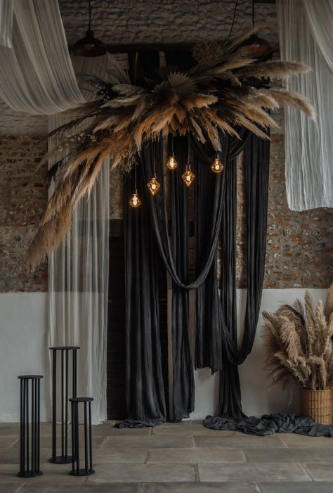 This image shows a barn wedding ceremony area. The main focus is on a hanging dried floral installation. The installation consists of normal cream pampas leaves and dark black ones, creating a depth in the installation. There are also uniquely shaped light bulbs hanging from the installation. Behind the installation are black and white drapes hanging across the venue ceiling and down the wall. Below the flowers are black plinths. Black Wedding Drapery, Black Wedding Draping, Suspended Floral Arrangements, Fabric Hanging From Ceiling, Yalda Decor, Backdrop Ideas For Wedding, Ceiling Installation Wedding, Diy Ceremony Backdrop, Hanging Flowers From Ceiling