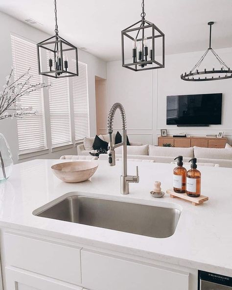 A modern kitchen features an island with white cabinetry and a white quartz countertop fitted with a stainless steel sink and gooseneck faucet. Branches with white flowers are arranged in a clear vase resting on the countertop, and amber soap bottles are placed on a bamboo tray next to the sink. Windows fitted with blinds fills the room with soft, filtered, natural light, and two black lantern pendant lights are hung over the island...   Image: theprettysocialcollective Sink On Island, Kitchen Island With Sink Decor, Sink In Island Kitchen, Island With Sink, Island Sink, Kitchen Sink Countertop, Warm Wood Flooring, Quartz Sink, Sink In Island