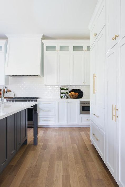 Stacked white shaker cabinets adorned with brass hardware and fixed beside a white wood paneled refrigerator located facing a gray island topped with a white quartz countertop. White Wood Paneling, Beautiful White Kitchens, Two Tone Kitchen Cabinets, White Oak Hardwood Floors, White Shaker Cabinets, Two Tone Kitchen, White Subway Tiles, Oak Hardwood Flooring, White Marble Countertops