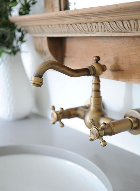 An antique brass vintage style wall mount faucet with cross handle knobs is mounted to a beneath a French wooden vanity mirror over a sink fitted to a gray quartz washstand top. Antique Bathroom Fixtures, Vintage Faucet Kitchen, Vintage Faucets Bathroom, Wall Mount Vanity Faucet, Antique Washstand Vanity, Wall Mounted Sink Faucet, Faucet Coming Out Of Wall, Antique Brass Vanity Faucet, Wall Mount Sink Faucet