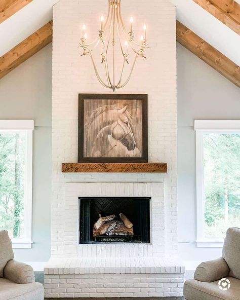 White painted brick floor to ceiling fireplace features a dark wood frame horse painting perched on the stained wood mantel. The fireplace is lit by a beige chandelier in this living room boasting white vaulted ceilings with wood beams. White Brick Fireplace Vaulted Ceiling, Above Fireplace Ideas, Shiplap Living Room, Faux Fireplaces, Stained Wood Beams, Grey Stone Fireplace, Two Story Fireplace, Fireplace Facing, White Brick Fireplace