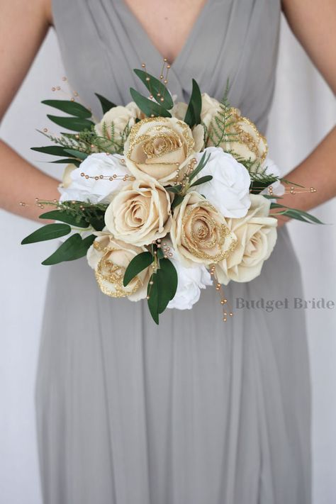 This lovely assortment of gold glitter tip, champagne and white roses and luxurious greenery is bound to turn heads on your wedding day. This lovely cascading brides’ bouquet from the Revy collection is an absolutely breathtaking arrangement for any bride to be, in any season. Gold leaves and gold pearls complete the look and add a delicate realism to this beautiful bouquet. This bouquet is 10” wide, 18” long, and is crafted by our in-house florists without using a foam base to anchor the flower Bouquets For Weddings Brides, White Green And Gold Wedding Bouquet, Sage And Gold Bouquet, Bridal Bouquet White And Champagne, Champagne Gold Flowers, Cream And Gold Bouquet, Cream Gold And Green Wedding, White And Gold Prom Bouquet, Sage Green And Gold Bouquet