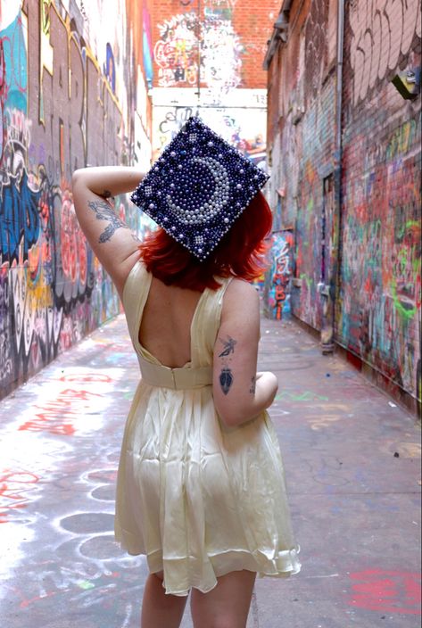 College senior graduation photo with beaded celestial graduation cap design inside alley filled with graffiti Gad Cap Ideas, Star Grad Cap Ideas, Sun And Moon Graduation Cap, Lana Del Ray Graduation Cap, Celestial Graduation Cap, Astronomy Graduation Cap, Artistic Graduation Cap, Witchy Graduation Cap, Physics Graduation Cap