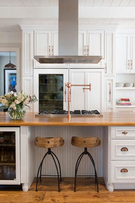 Stunning kitchen features white raised panel cabinets adorned with copper hardware framing a glass door refrigerator. Farmhouse Kitchen Pot Filler, Island Pot Filler, Stove In Island With Hood, Island Kitchen Hood, Kitchen Island Hood Vent, Stove In Kitchen Island, Island Hood Vent Ideas, Kitchen Island Stove, Kitchen Island Hood