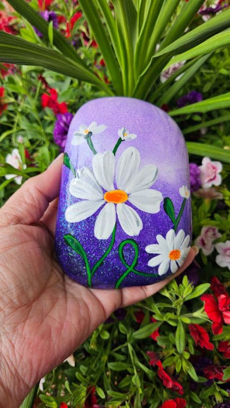 Painted River Rocks.  Hand painted daisies over a sparkling gradient colored background.  Very pretty!   Three color options.  Purple, Blue Teal or Green Teal.  Wonderful gift.  Home/Office Décor.  These look amazing in a flower pot or just on a shelf.  They do not stand up on their own but easily lean or can just lay flat on a coffee table.  Sealed in clear gloss resin for permanent protection and shine.  If outdoors, try to place in more shade than sun to prevent potential color fade.  Bring indoors over winter.  Backsides are blank with my signature.  Price does include $11 Priority Mail shipping. Purple Painted Rocks Ideas, Rock Painting Ideas Flowers, Flower Rocks, Summer Rocks, Painted Daisies, Rock Painting Flowers, Painted River Rocks, Rock Flowers, Acrylic Painting Flowers