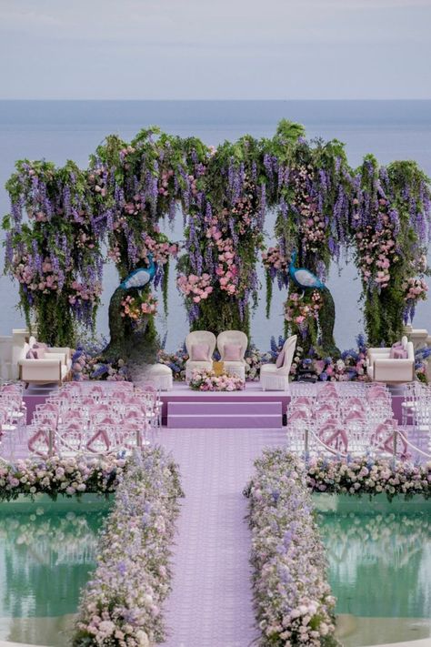 This dreamy oceanfront wedding setup features a stunning lavender aisle leading to a grand floral backdrop with cascading wisteria and pastel blooms. The intricate floral arrangements are complemented by clear seating with lavender accents, creating a truly romantic and elegant atmosphere. Perfect for couples seeking a luxurious and unique outdoor wedding by the sea. #OceanfrontWedding #LavenderWedding #FloralBackdrop Florals On Arch, Unique Ceremony Seating, Flower Field Wedding Ceremony, Blue Florals Wedding, Lavender Backdrop, Unique Outdoor Wedding, Wedding Themes Outdoor, Weddings 2024, Wedding Aisles