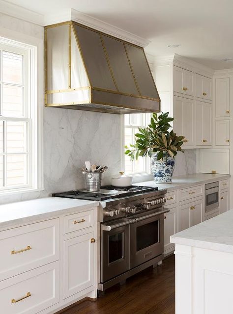 A steel hood accented with a brass trim is fixed between windows to a honed white marble backsplash above a stainless steel dual range flanked by white shaker cabinets adorning brass hardware and a honed white marble countertop. White Marble Backsplash, Shaker Style Kitchen Cabinets, Kitchen Hood Design, Kitchen Cabinet Trends, Replacing Kitchen Countertops, Kitchen Hood, Shaker Style Kitchens, Kitchen Cabinet Styles, Kitchen Hoods