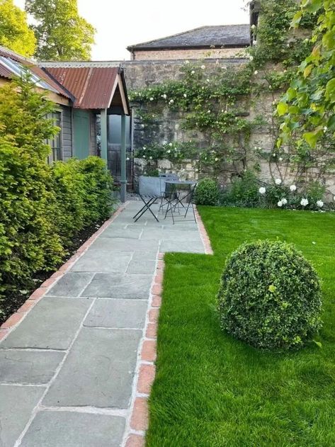 light grey path to the left edged with red brick. green grass and topiary bush right. Stone wall at top of image covered in green climber plants with white flowers and a green shed with a brown corrugated iron roof to the left with a table and chairs outside. Paving Pathway, Flower Shed, Paving Design, Beautiful Home Gardens, Brick Detail, Brick Pavers, The Brick, Garden Paths, Westminster