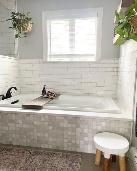 Gray grid tiled white drop-in bathtub with black faucet features a glazed white staggered tile tub surround. A dark wood bath tray is placed across the tub under a white bathroom window with white blinds. Shower Tub Window, Tubs With Windows, Bathtub With Surround, Tile Around Drop In Tub Master Bath, White Tile Around Bathtub, Bathtub With Tile Surround Ideas, Tile Around Master Bathtub, Built In Bathtub Tub Surround Tile, Drop In Tub With Tile Surround