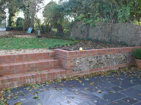 Slate patio with brick and flint retaining wall. Brick And Flint Garden Wall, Red Brick Retaining Wall Ideas, Red Brick Retaining Wall, Courtyard Flooring, Flint Wall, Circle Patio, Brick Wall Gardens, Slate Patio, Garden Retaining Wall