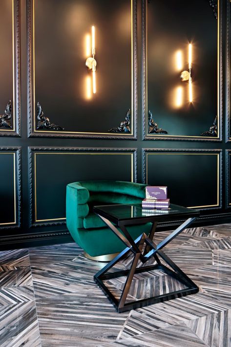 Black wall panelling with gold trim and corner embellishments adds texture and a sophisticated base for this sitting area design. Wand sconce lighting is mounted within the panels reflecting a warm glow on the black paint. The deep, vivid green of the velvet chair looks beautiful against the wall and black and white patterned floor. A geometric table adds the finishing touch to the sleek concept. Art Deco Wall Panelling, Wall Panelling Ideas, Sitting Area Design, Panelling Ideas, Green Velvet Chair, Art Deco Ideas, Trellis Wallpaper, Art Deco Interior Design, Wall Panelling