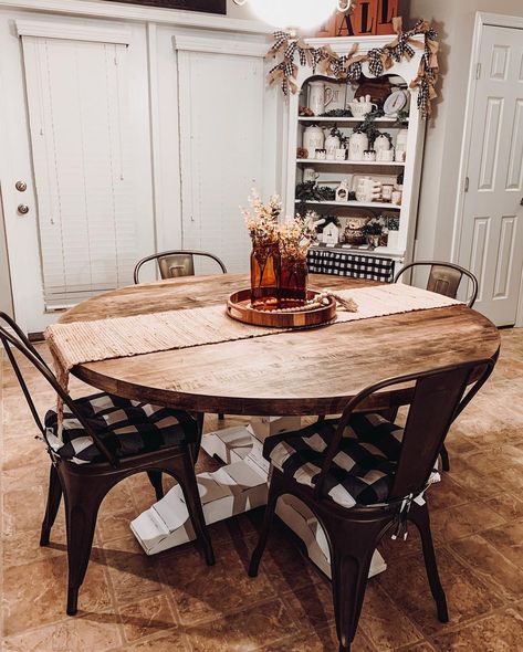 A white corner hutch stands in the corner of a farmhouse kitchen. A round dining table with a stained wood top and thick white base is centered on a brown vinyl floor. Black metal chairs with padded buffalo-checked cushions are arranged around it. A brown woven runner with a round tray and brown vases of dried flowers accentuate the table as a centerpiece. Wood Table Metal Chairs, Round Dining Table With Runner, Round Dining Table Centerpiece, Round Kitchen Table Decor, Round Farmhouse Dining Table, Table Runner Round Table, Dining Table Centerpiece Ideas, Farmhouse Round Dining Table, Round Table Runner