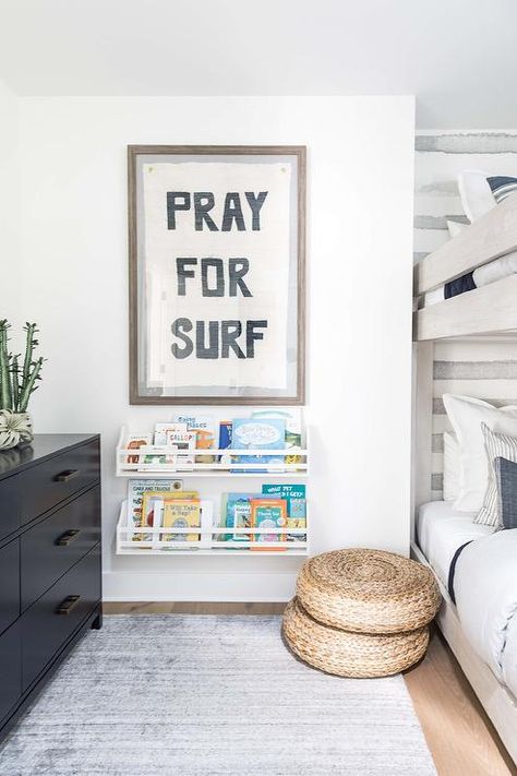 A 'Pray For Surf' art print hangs in a brown wooden frame over stacked white bookshelves fixed over seagrass poufs positioned in a gray boys' bedroom on a gray rug beside white weathered bunk beds. Gray Boys Room, Boys Beach Bedroom, Surf Interior, Salt Interiors, Surf Bedroom, Surfer Room, Pure Salt Interiors, Pray For Surf, Surf Room