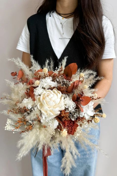 Your special day deserves a bouquet that reflects your free spirit and love for nature. This handcrafted boho bridal bouquet, bursting with warm terracotta, burnt orange, and sunshine yellow blooms, evokes the magic of autumn. Soft pampas grass adds a touch of texture, while luxurious cafe au lait roses whisper romance. Hand-tied with love, this bouquet is the perfect complement to your bohemian spirit and a forever memento of your rustic wedding celebration. Terracota Bridal Bouquet, Burnt Orange And Pampas Wedding Flowers, Fall Wedding Bouquets Boho, Dried Flower Arrangements Wedding, Burnt Orange Wedding Flowers, Dry Flower Bouquet, Boho Bridal Bouquet, Autumn Soft, Preserved Hydrangea