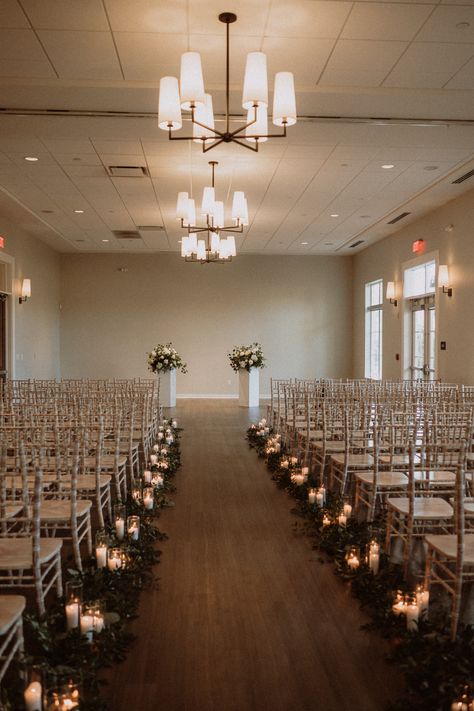 This photo shows an indoor wedding ceremony with greenery and clusters of pillar candles going down the length of the aisle. The ceremony chairs are white. The backdrop of the ceremony are two white columns with floral arrangements sitting on top. The fresh florals are white and have accents of greenery. Wedding Isle Candles, Candle Aisle Decor, Candle Aisle, Wedding Alter Flowers, Wedding Aisle Candles, Tea Light Candles Wedding, Taper Candles Wedding, Aisle Candles, Pillar Candles Wedding