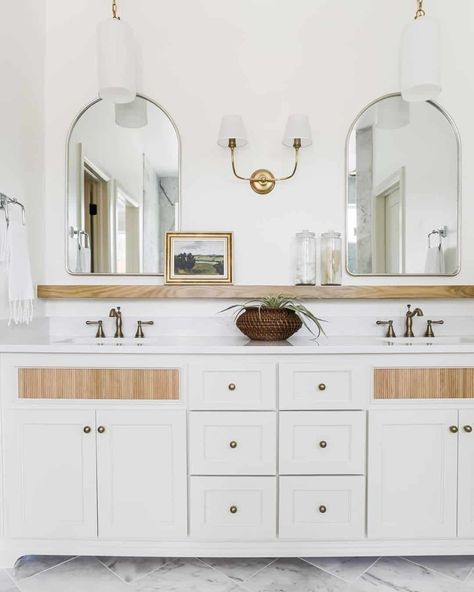 White and brass dual head sconce is flanked by silver frame arched bathroom mirrors mounted above a light wood floating shelf. A white dual sink vanity under the shelf is lit by white and brass pendant lights. Shelf Above Sink, Bathroom Mirror With Shelf, Bathroom Mirror Storage, Rectangular Bathroom Mirror, Vanity Shelves, Bathroom Sconces, Wood Floating Shelves, Mirror With Shelf, Storage Mirror