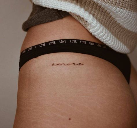 Tattoo of the word "amore" located on the hip. Amor Tattoo On Hip, Small Tattoos For Women Hip Bone, Amor Hip Tattoo, Words Hip Tattoo, Danty Tattoos Small On Hip, One Word Hip Tattoo, Upper Thigh Writing Tattoo, Amore Tattoos For Women, Hidden Hip Tattoo