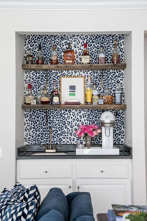 Black and white leopard print wallpaper accents a wet bar boasting brown wood floating shelves mounted above an antique brass faucet fitted to a black countertop accenting white cabinets. Wet Bar Nook, Alcove Bar, Wet Bar Ideas, Wet Bar Cabinets, White Leather Bar Stools, White Beveled Subway Tile, Bar Nook, Leopard Print Wallpaper, Light Gray Cabinets