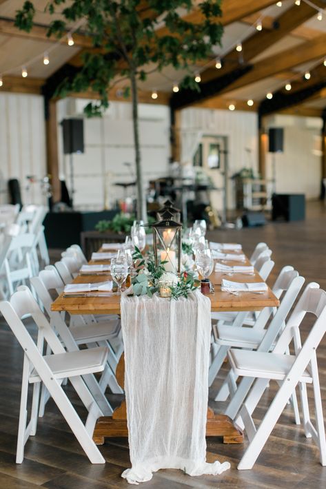 Wedding reception long wooden tables with white draped table runners, lanterns, and white red and greenery centerpieces with twinkle lights at wedding. #weddingreception #rusticweddingdecor #weddingdecor #weddingtablescape White Table Runners Wedding, Wood Tables With White Chairs Wedding, Lantern Centerpieces Long Table, Wooden Tables Wedding, Wooden Table Wedding Decor, Lights At Wedding, Lobola Decor, White Chairs Wedding, Lantern Table Centerpieces