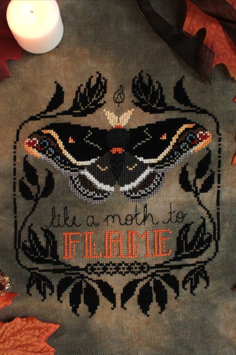 A finished cross stitch pattern of a black moth with orange accents. A stylized black leaf border surrounds the moth. Cursive text reading "like a moth to" is backstitched underneath. The word "flame" is in all caps and is orange. Moth Cross Stitch, Halloween Moth, Gothic Cross Stitch, Stitch Stuff, Stitching Projects, Graph Patterns, Gothic Cross, Gothic Crosses, Leaf Border