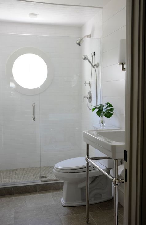 Sleek bathroom features 2-leg washstand with white porcelain sink next to toilet and shower with seamless glass door, white subway tile surround, round window and taupe tiled shower floor. Windows In Bathroom, Taupe Bathroom Ideas, Benjamin Moore Moonshine, Greige Bathroom Ideas, Greige Bathroom, Taupe Bathroom, White Subway Tile Shower, Zen Bathroom Decor, Window Bathroom