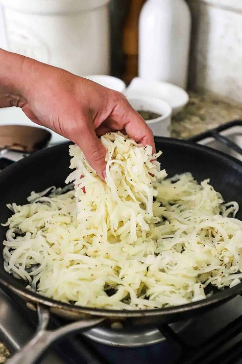 A person dropping a handful of shredded partially cooked russets potatoes in a large non-stick skillet on a stove. Best Way To Cook Frozen Shredded Hashbrowns, How To Cook Frozen Hashbrowns In Skillet, Homemade Hashbrowns Shredded, Frozen Shredded Hashbrown Recipes, Shredded Breakfast Potatoes, Skillet Hashbrowns, Homemade Home Fries, Fried Shredded Potatoes, Shredded Hashbrown Recipes