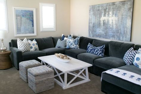 Amazing living room features a a blue and grey abstract art piece placed above a dark grey velvet sectional adorned with an assortment of blue pillows facing a white trestle coffee table and a pair of blue and grey cube poufs. Grey Couch Blue Rug, Blue Grey Sectional, Relaxed Living Room Decor, Blue Transitional Living Room, Charcoal Living Room, Grey Sectional Couch, Gray Sectional Living Room, Dark Grey Living Room, Relaxing Room