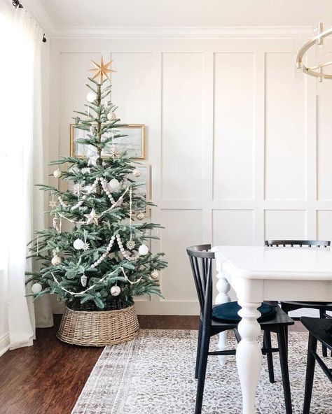 A Christmas tree with silver and white ornaments stands in a tree collar in front of a white board and battened dining room wall. A gold chandelier hangs over the white dining room table, while black chairs are placed on a patterned rug laying on the dark wood floor...   Image: caitlinmariedesign Noble Fir Christmas Tree, Neutral Christmas Tree, Minimalist Holiday Decor, Best Artificial Christmas Trees, Neutral Holiday Decor, First Christmas Together Ornament, Minimalist Christmas Decor, Fir Christmas Tree, Holiday Mantel