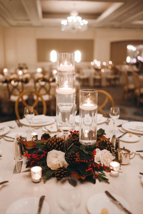 A wedding table setting with white tablecloths, candles, pine cones, red berries, and white flowers. Christmas Centerpieces For Weddings, December Wedding Decorations Wedding Table Decor, Christmas Decor Ideas Wedding, Christmas Wedding Tablescape, Christmas Wedding Round Table Decor, Simple Winter Wedding Table Decor, Christmas Sweetheart Table Wedding, Winter Branch Centerpieces, December Wedding Centerpieces